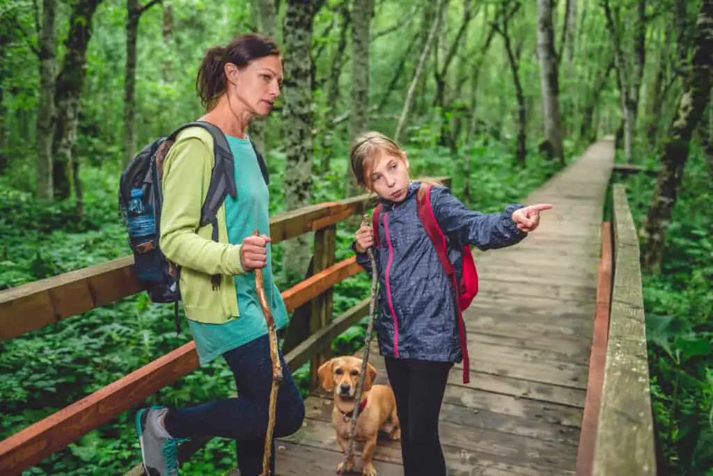 woman and girl hiking across bridge with their dog with filled backpacks