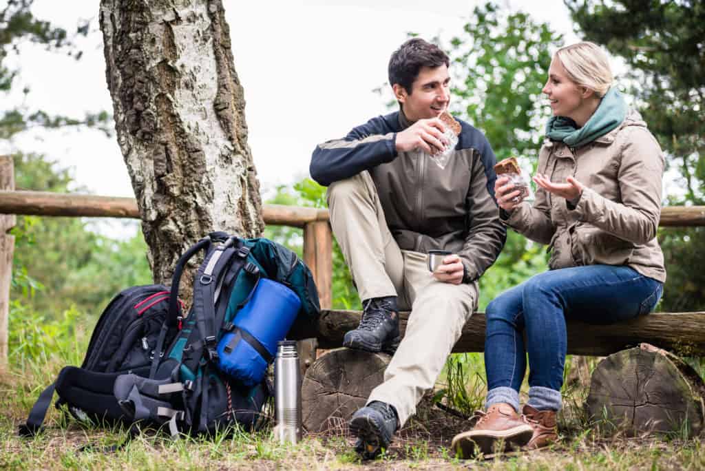 couple eating sandwiches sitting on logs, their backpacks filled with day hiking gear next to them