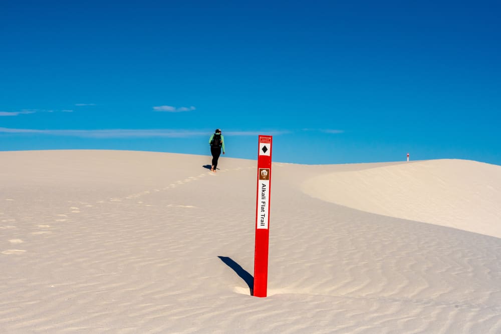 hiker walking across sand dunes with sign in front for the alkali flat trail