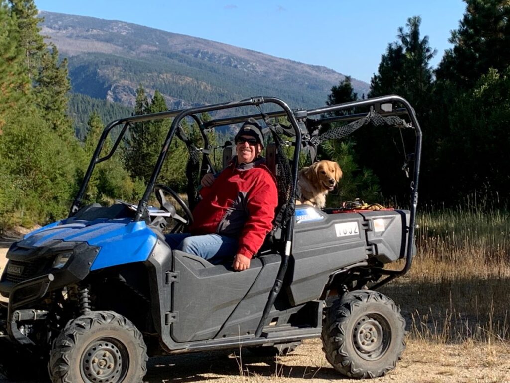 Man in red jacket with golden retriever riding in a side by side in mountains