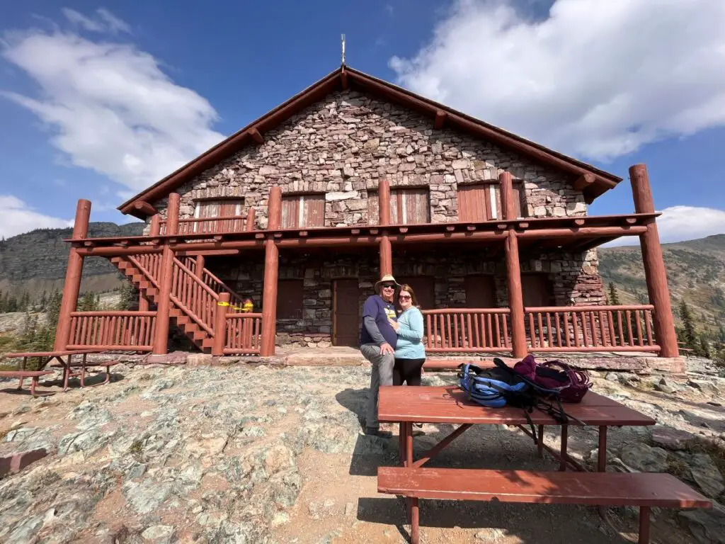 man and woman standing in front of stone Granite Park chalet with wooden rails on mountain top