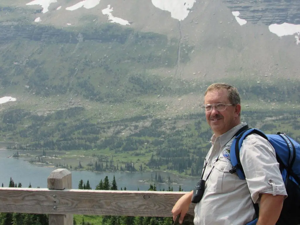 Man standing against a wooden railing with mountains in the background. Hidden Lake Overlook