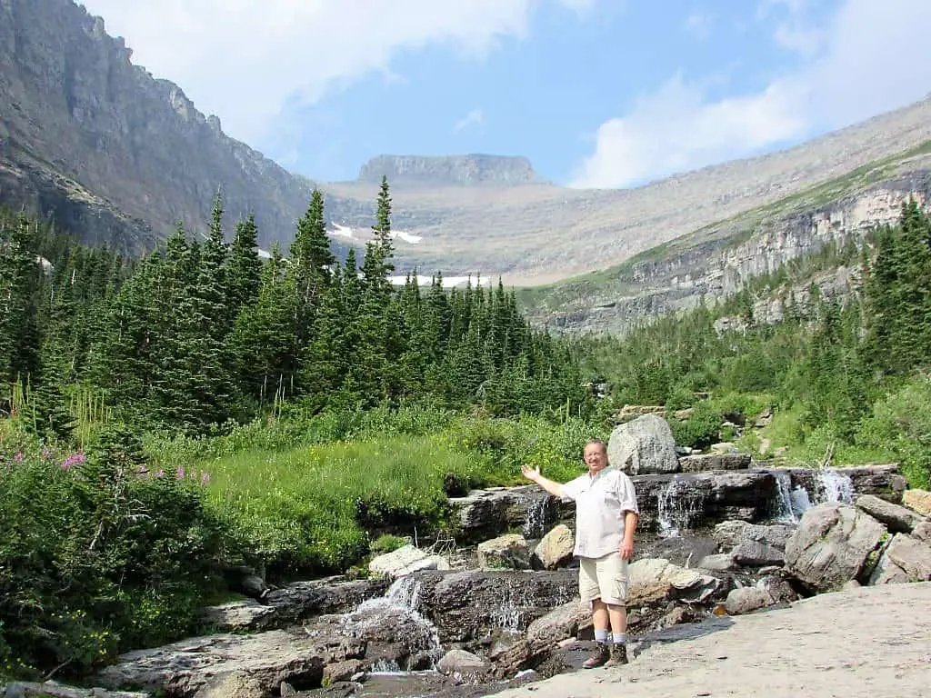 Man standing in front of water cascading over boulders with mountain valley in the background. Just off Going to the Sun Road at Lunch Creek