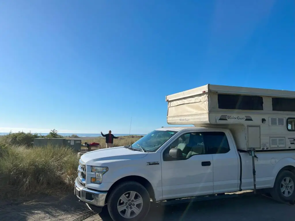 truck camper in front with beach in background. Man waving. Blue sky. Campground in Gold Bluffs Beach part of the REdwood National park itinerary