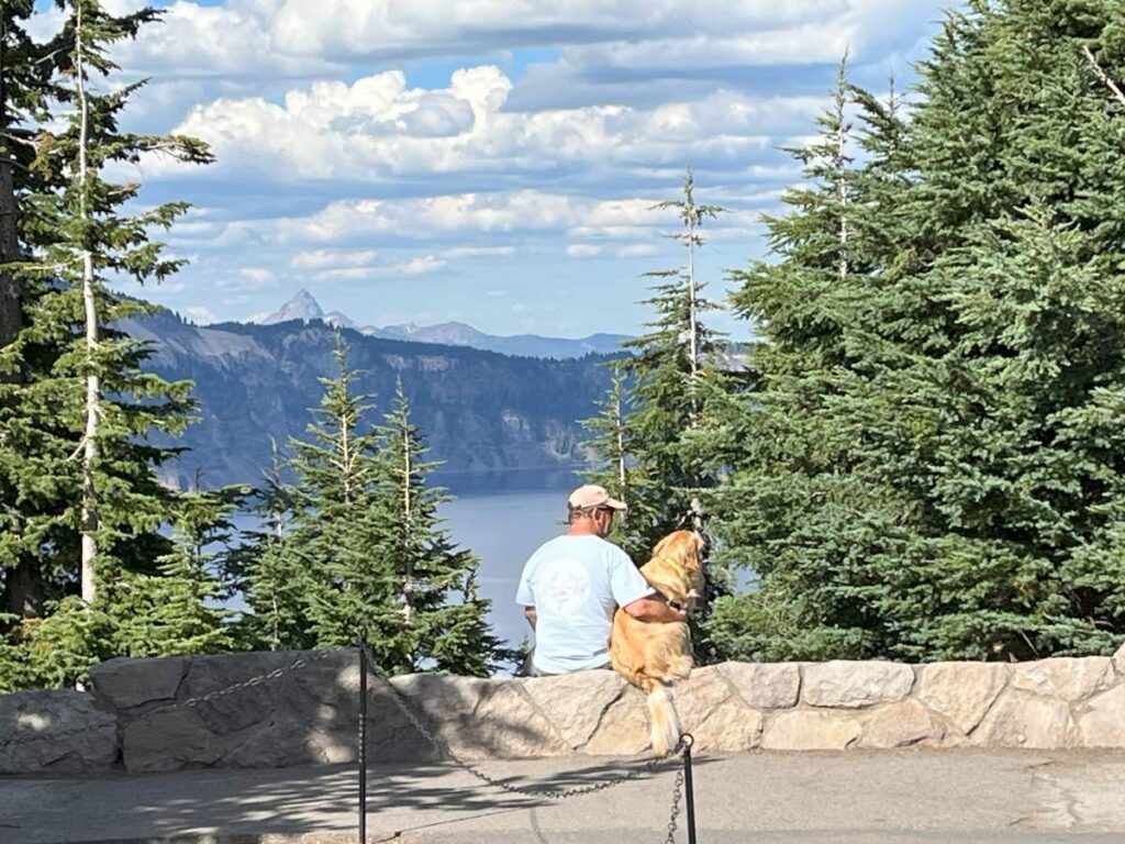 man and dog sitting on stone wall with crater lake in background