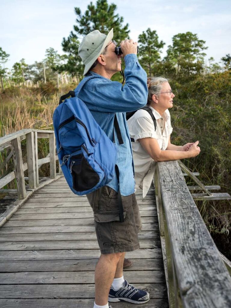 Man and woman standing on a wooden bridge watching birds through binoculars